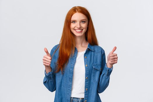 Waist-up portrait alluring confident smiling redhead female blogger review awesome new product, showing thumbs-up in approval, like or agree gesture, standing white background.