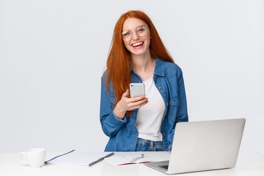 Cheerful, friendly-looking charismatic redhead female designer, freelancer with red foxy hair, glasses, laughing carefree, holding smartphone, working over project with laptop, drink coffee.