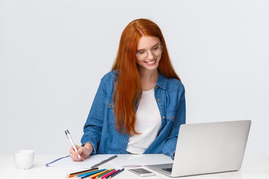 Education, work and vanlife concept. Cheerful lovely redhead woman in glasses standing near table, reading something from laptop, gather information from internet to write good essay.