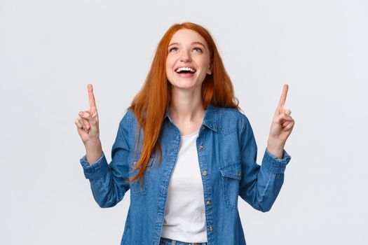 Cheerful charismatic redhead woman in denim shirt, looking and pointing up with happy smile, laughing, found exactly what need, dreaming, standing white background positive emotions.
