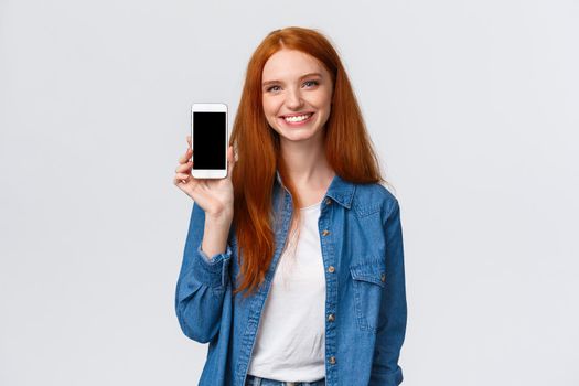 Cheerful friendly redhead woman checking water app, stay hydrated with smartphone application, showing mobile screen and smiling delighted, standing white background in casual clothes.