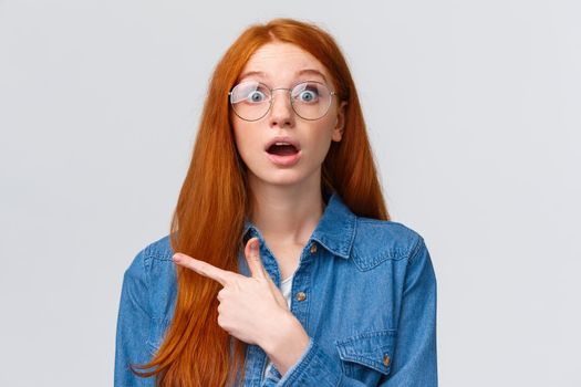 Surprised and wondered, amused attractive redhead woman in glasses, red natural hair, freckles, blue eyes open mouth fascinated and curious, pointing finger left, asking question about banner.