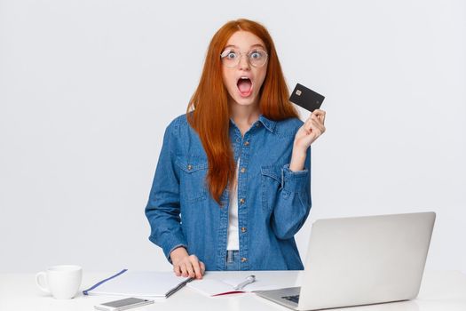 Excited and shook, impressed redhead girl holding credit card, got cashback for purchasing new furniture online store, visit website good prices, stand white background with laptop.