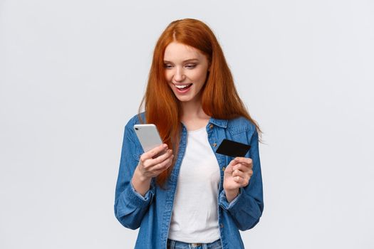 Shopaholic, internet concept. Waist-up portrait cheerful friendly redhead woman order online, insert bank info register on site, taxi order application, hold credit card and smartphone, smiling.