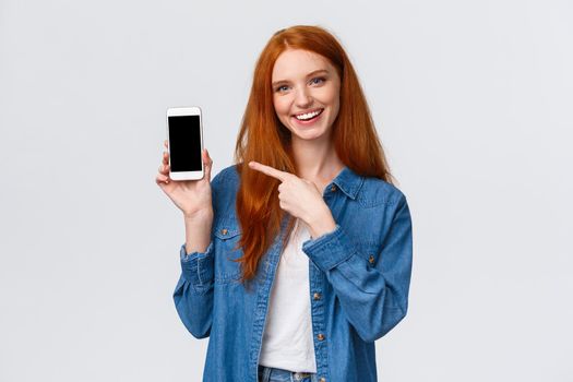 Waist-up portrait joyful charismatic redhead female review smartphone application, showing her photo with cute filter, pointing mobile display and smiling camera, white background.