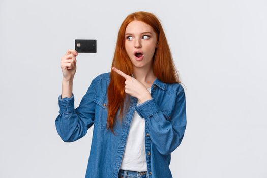 Discounts, shopping and banking concept. Waist-up impressed, thrilled cute redhead girl in denim shirt holding credit card, pointing at it with fascinated impressed eyes, standing white background.