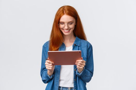 Waist-up portrait pretty happy redhead female student, studying, working over design project, wearing glasses, holding digital tablet and looking satisfied gadget screen, smiling pleased.