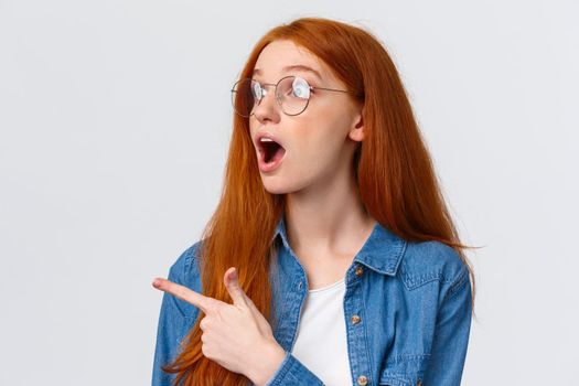 Cool take a look. Close-up portrait fascinated and excited cute redhead woman in glasses checking out awesome banner, pointing and looking left with opened mouth, say wow, white background.