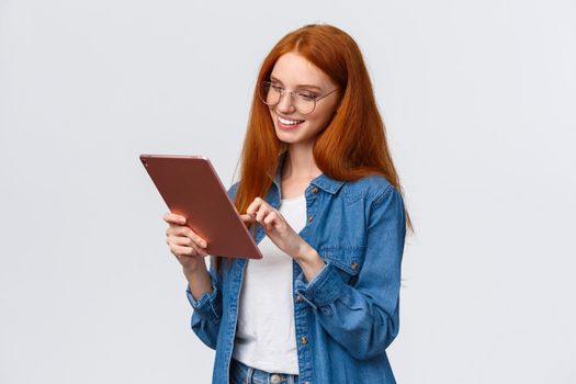 Waist-up portrait carefree european woman with red hair in glasses, looking entertained and amused at digital tablet, smiling delighted, pleased with result, standing white background.