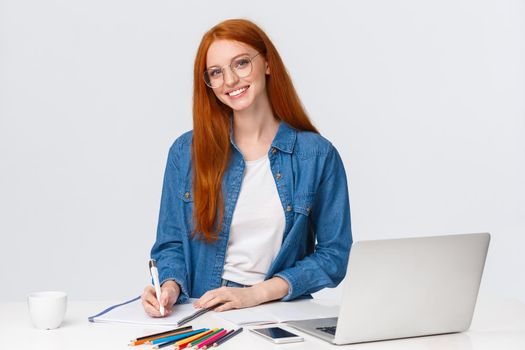 Talented creative female designer working over new project, prepare drawings for art class, standing near table, taking notes, use laptop and colored pencils, white background.