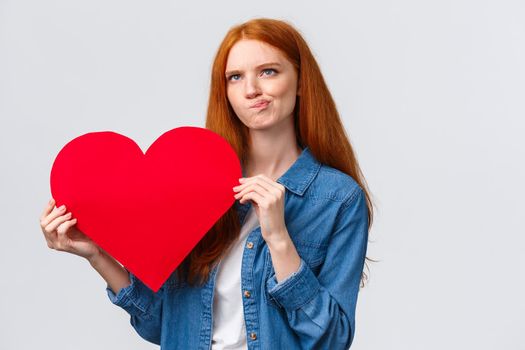 Thoughtful and focused, serious-looking redhead girl trying think, smirk indecisive and unsure, looking up pondering, holding big red valentines day heart, white background.