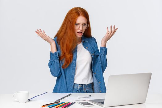Mad and confused, frustrated redhead girl having troubles with computer, working on hard project, raising hands up dismay and annoyance, shrugging, stare laptop screen bothered.