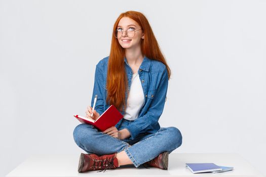 Dreamy and creative cute diligent female student with red hair, in glasses, sitting on floor with legs crossed surrounded by notebooks writing something, creating poem, look up thoughtful.
