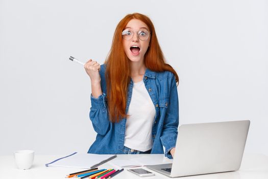 Amused, excited and surprised cute redhead girl telling roommate awesome news, read online great event nearby, smiling scream fascinated, standing near laptop and table with colored pencils.