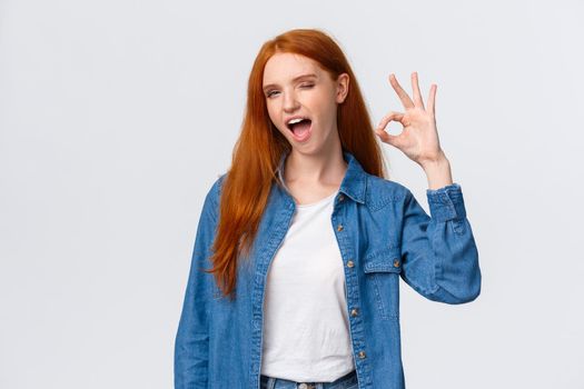 Waist-up portrait cheeky, confident and relaxed, chill redhead woman saying all alright, encourage everything okay, showing ok gesture wink and tongue, standing white background.