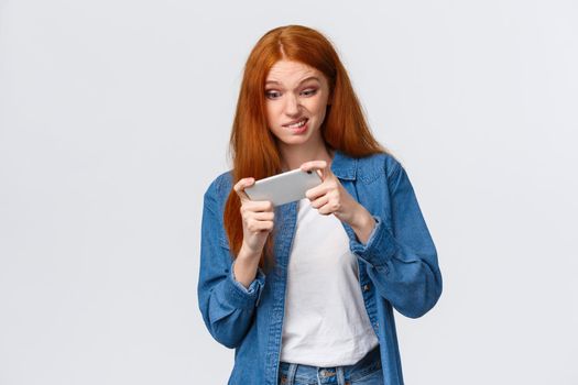 Girl having intense fighting round in game, tap screen and trying beat friends score in app. Excited good-looking redhead female student wasting time playing smartphone arcade, white background.