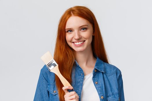 Close-up portrait talented and creative cute redhead girl with blue eyes like drawing, attend art courses, holding painting brush, consider paint walls new color, smiling confident, white background.