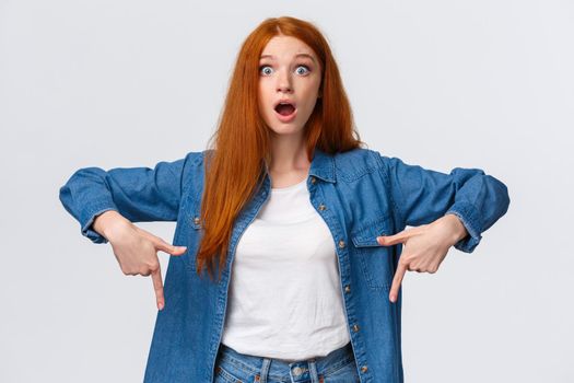 Amazing, have you heard already. Astonished, speechless and impressed redhead woman in denim shirt, drop jaw, gasping stare camera fascinated, say wow, pointing fingers down.