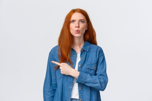 What is that. Alluring redhead girl in denim shirt, staring questioned, asking question and pointing finger left, frowning perplexed, dont know something, curious about product, white background.