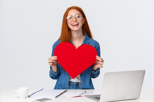 Valentines day, creativity and feelings concept. Cheerful smiling redhead girl with long-distance relatioship sending her love through internet, using web cam to show big red heart and say love you.