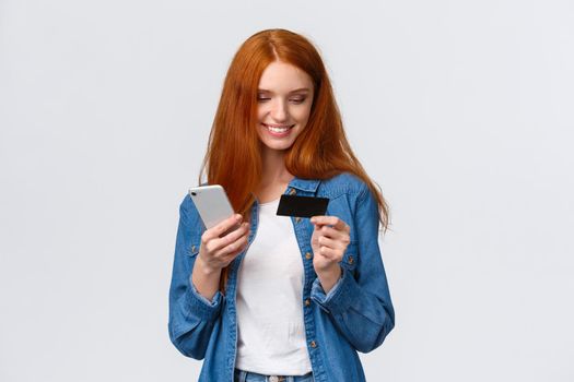 Finance, business and education concept. Cheerful cute alluring redhead woman making online purchase, holding smartphone and credit card, smiling as buying present for valentines day internet store.