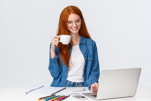 Lovely good-looking redhead female working on new project in office, standing near table with laptop, looking at device screen, smiling drinking coffee, having break from work, white background.