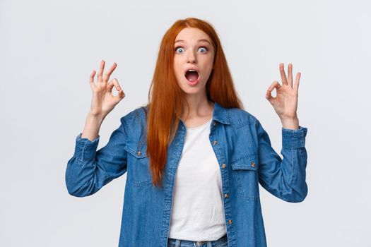 Amused, excited and thrilled good-looking redhead female in denim shirt, open mouth, drop jaw speechless staring with popped eyes camera, showing okay gesture in approval, white background.