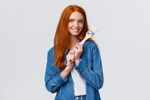 Creative pretty redhead female in denim shirt, creating new art, painting, making repair works in own room, holding paintbrush and smiling tempting, standing white background.