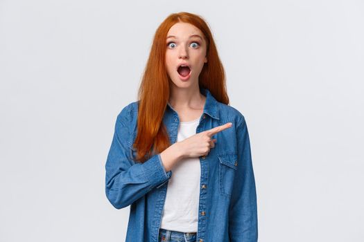 Have you seen it, awesome news. Waist-up portrait amazed, impressed redhead female in denim casual shirt, drop jaw, gasping and stare astonished as pointing finger right, white background.