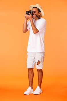 Full-length vertical shot african american male tourist taking pictures of sightseeing, looking in camera lense to take perfect shot, photographing girlfriend on beach, standing orange background.