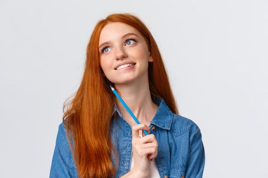 Lovely and creative, thoughtful dreamy smiling redhead woman looking up and thinking, imaging something beautiful, holding colored pencil, drawing, attend art courses, white background.