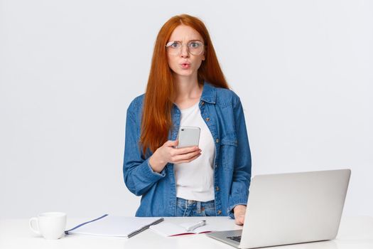 Disturbed, puzzled and confused serious-looking woman with red hair in glasses, staring perplexed camera, read strange worrying message on smartphone while working on project with laptop.