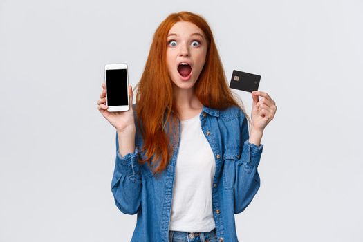 Waist-up portrait excited, astonished and thrilled redhead female student holding credit card, showing smartphone display, application, drop jaw impressed, looking camera astounded, white background.