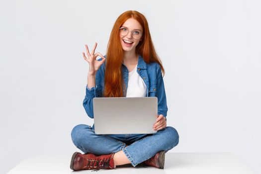 Dont worry, all done, everything under control. Confident happy and relaxed redhead girl saying all okay, show ok gesture while sit on floor with legs crossed, holding laptop, white background.