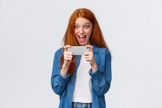 Excited, happy and surprised pretty redhead female student beat score, won competition, challenge in smartphone game, holding phone horizontally, watch funny video from party, look astonished display.