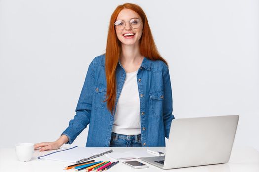 Education, work and freelance concept. Attractive charismatic redhead female digital nomad, designer working remote, creating design project, standing near table with laptop, colored pencils.