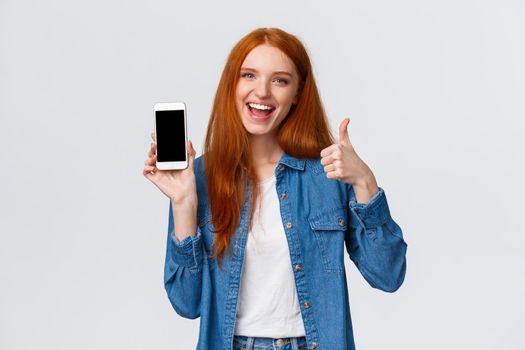 Girl showing friend cool new mobile game. Attractive cheerful redhead woman holding smartphone, introduce application, telephone app, make thumb-up and smiling in approval, recommending.