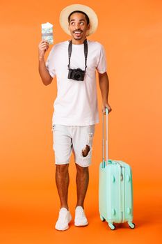 Full-length vertical shot excited happy african american guy dreaming about holiday, looking up amused and imaging travel, holding passport and flight tickets, luggage, orange background.