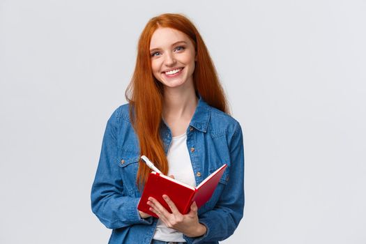 Waist-up portrait lovely redhead teenage girl taking notes during favorite college class, holding pen and red notebook, smiling carefree camera, writing something, make to-do list.