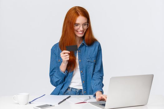 Pleased, satisfied relaxed cute redhead woman making order from work, paying online, insert credit card number billing info, using laptop to purchase, standing white background.
