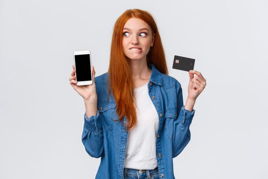 Shopaholic cant hold herself want buy something online. Eager and excited redhead girl see tempting price internet store, biting lip thrilled stare smartphone display, hold credit card.