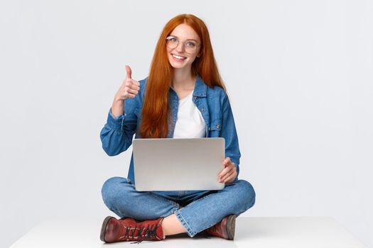 Education, internet and people concept. Female redhead digital nomad, freelance girl in glasses, finished project before deadline, sitting on floor with crossed legs and laptop, show thumb up.