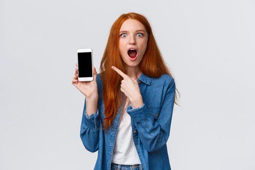 Shocked young woman saw her ex-boyfriend with someone else on internet social media. Astonished and impressed redhead woman gossiping pointing at smartphone display, drop jaw.