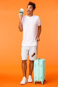 Full-length vertical shot dreamy, excited african-american guy on vacation, holding luggage, carry suitcase and passport with flight tickets, imaging perfect trip, awesome journey, orange background.
