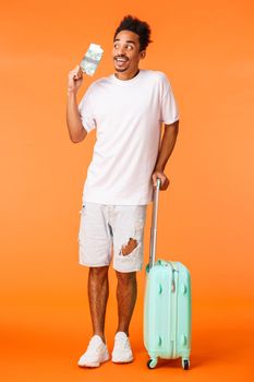 Full-length vertical shot excited dreamy and happy young african-american man, imaging perfect vacation, holding luggage and passport with flight tickets, travelling, orange background.