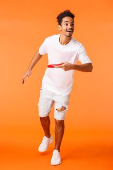 Full-length vertical shot happy, charismatic african-american sporty guy like active games outdoors, throwing frisbee at friend and smiling, enjoying beautiful sunny summer day, orange background.
