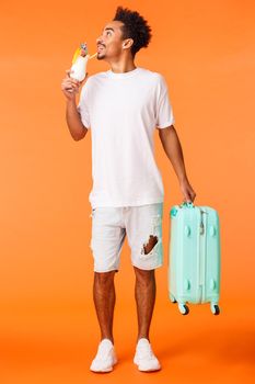 Full-length vertical shot dreamy happy and relaxed african-american man carrying luggage, suitcase and drinking cocktail, looking around pleased and smiling, book in luxury hotel, orange background.