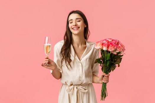 Celebration, party and beauty concept. Romantic beautiful young woman in dress, holding flowers enjoying celebrating, drinking champagne from glass, close eyes and laughing, pink background.