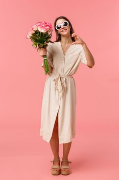 Full-length vertical portrait romantic and sensual, coquettish young woman holding bouquet flowers and make heart sign, expressing affection being touched and grateful with roses, pink background.