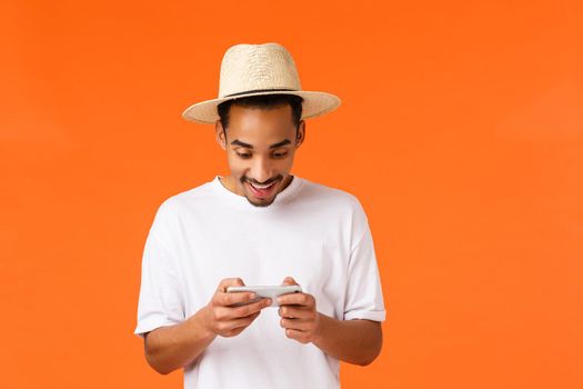 Amused geeky african-american guy playing smartphone game in airport while waiting for flight, holding mobile horizontally entertained, download racing app, tap screen, want win, orange background.